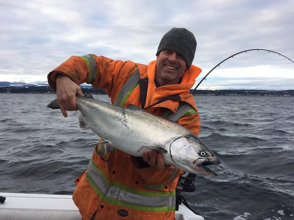 A man proudly presents a fish he caught, he is on the deck of a boat and is smiling at the camera, wearing an orange, high-visibility jacket. Calm waters and cloudy skies are visible in the background.